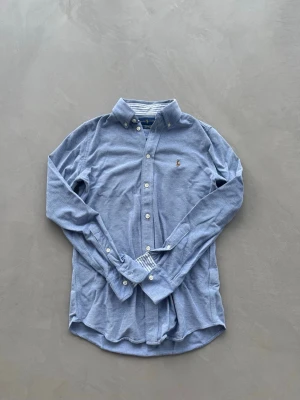 Blå skjorta från Polo Ralph Lauren - OBS! KOMMER STRYKAS INNAN FRAKTAT! Klassisk blå skjorta från Polo Ralph Lauren med button-down krage och broderad logga på bröstet. Skjortan har långa ärmar, vita knappar och är tillverkad i mjuk bomull. Perfekt till jeans eller chinos för en clean look. Passar dem runt 160-170