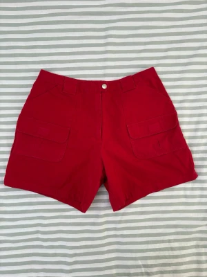 Vero Moda röda shorts - Röda shorts med hög midja och fickor fram och bak. Shortsen har en knapp med texten ”Vero Moda” samt kardborreförslutningar på både fram- och bakfickorna. Supergulliga och figursmickrande shorts