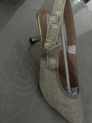 ENDAST  🌸~1000kr🌸 - Säljer ett par eleganta pumps från Christian Dior i beige tyg med broderade blommönster. Skorna har spetsig tå, öppen häl med justerbar rem och en smal guldfärgad klack. Perfekta för dig som vill ha en lyxig och trendig look. helt nya oanvända, tyvärr passar dom ej! A K0PIOR💕FINNS I HÖGKLACK OCKSÅ!