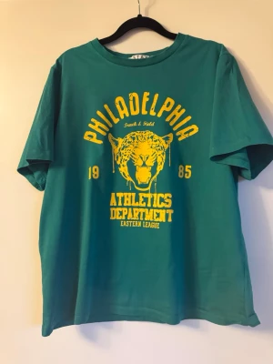 Grön Philadelphia Athletics T-shirt - Cool grön t-shirt från STAY med stort gult tryck framtill: 'Philadelphia Athletics Department' och en tiger. T-shirten har rund hals, korta ärmar och är i mjuk bomull.