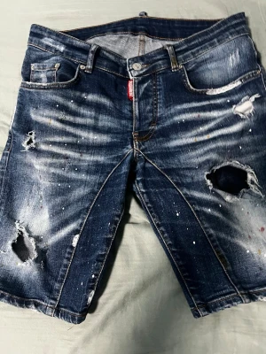 Blå slitna jeansshorts med hål - Säljer ett par blå jeansshorts med slitningar, hål och färgstänk för en riktigt street och avslappnad look. Klassisk femficksmodell med knappgylf och råa kanter. Perfekta för dig som gillar en edgy stil.