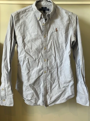 Ljusblå slim fit skjorta Ralph Lauren - Klassisk ljusblå skjorta från Ralph Lauren i slim fit-modell. Skjortan har långa ärmar, knappar framtill och den ikoniska broderade loggan på bröstet. Tillverkad i bomull och passar perfekt till både jeans och chinos.