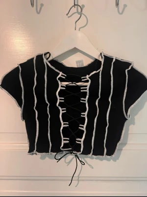 Svart croppad topp med snörning - Trendig svart croppad topp med vita vågiga sömmar och snörning framtill. Kortärmad modell med tight passform och coola detaljer som ger en edgy vibe. Perfekt för dig som vill sticka ut med din stil.