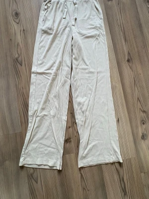 Beige ribbade mjukisbyxor med dragsko - Säljer ett par beige mjukisbyxor med ribbad struktur och raka ben. Byxorna har resår i midjan med dragsko och två sidofickor. Perfekta för chill dagar hemma eller när du vill vara bekväm men ändå se snygg ut.