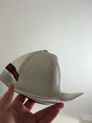 ÄKTA KEPS FRÅN GUCCI MODDEL 392’6 2003  - Säljer en vit keps från Gucci med diskret GG-monogram i tyget och klassiska gröna och röda ränder på sidan. Kepsen har böjd skärm och insida med svart kantband där Gucci-loggan syns. Materialet känns lyxigt och kepsen har en clean look.