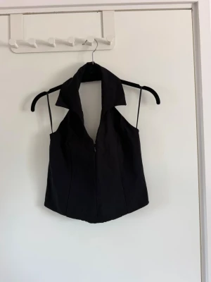 Svart ärmlös linnetopp med krage - Snygg svart linnetopp med bred krage och dragkedja framtill. Toppen är ärmlös med smala axelband och har en figurnära passform som ger en cool och trendig look. Perfekt att styla med jeans eller kjol för en modern vibe. Använd två gånger