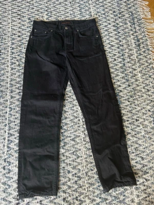 Svarta ”gritty jackson ” nudie jeans-31/32 - Ett par svarta nudie byxor som köptes i för liten storlek och därav säljs. Passformen heter Gritty Jackson är en rakare modell. Skick 9,5/10, lappen gick sönder lite i tvätten som kan ses på bild tre. Använda runt 5 gånger. Storlek 31/32. Nypris 1600