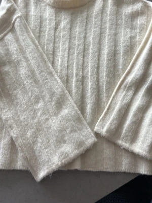 Vit ribbstickad tröja - Mysig vit stickad tröja med ribbade ränder och långa ärmar. Tröjan har en mjuk och fluffig känsla, perfekt för kyligare dagar. Enkel och stilren design som passar till det mesta. En liten och väldigt ljus fläck på framsidan längst ner. Och en liten skuggning på baksida ärm. 