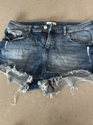 Blå jeansshorts med fransig kant - Snygga blå jeansshorts med slitna detaljer och fransig, rå kant. Klassisk femficksmodell med knapp och dragkedja framtill. Perfekta för sommaren och ger en avslappnad streetstyle-look.