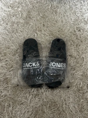 Slides från Jack & Jones *41* - Snygga  slides från Jack & Jones med vit logga över foten. Sköna att glida runt i tack vare den mjuka sulan och bred rem. Perfekta för chill dagar eller när du vill ha en enkel och stilren look. Tillverkade i syntetmaterial.