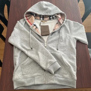 Burberry Zip Up Hoodie - Säljer en stilren grå hoodie från Burberry med klassiskt rutigt mönster i huvan. Använd 2 gånger då jag inte gillade den.