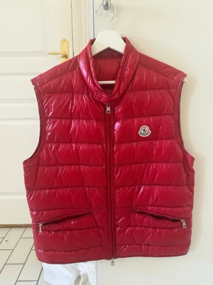 Röd dunväst från Moncler - Snygg röd dunväst från Moncler med glansig finish och klassisk logga på bröstet. Västen har två dragkedjeförsedda fickor och hög krage. Perfekt för lager på lager och ger en sportig vibe till din outfit. Passar en medium. Knappt använd då de inte riktigt va min killes stil. Storlek 3 motsvarar M men passar min kille som bruka ha S/m