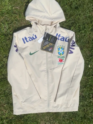 Vit Brasil Nike vindjacka med huva - Snygg vit vindjacka från Nike med huva och dragkedja. Jackan har Brasilien CBF-logga, grönt Nike-märke och tryck i blått och grönt på bröst och ärmar. Perfekt för dig som gillar fotboll och sportig stil.