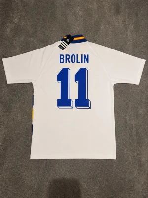Svensk landslagströja Brolin #11 Adidas - Klassisk vit svensk landslagströja från Adidas med Brolin och nummer 11 på ryggen. Tröjan har blå och gula detaljer, V-ringad krage med blå och gula ränder samt Sveriges landslagsemblem på bröstet. Perfekt för dig som älskar fotboll och retrostil.