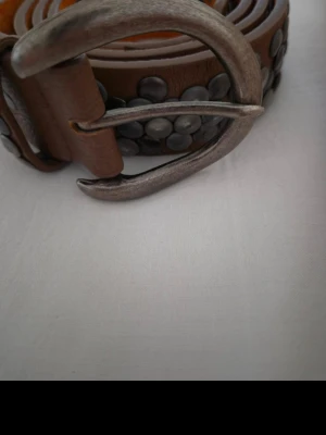 Brunt skärp med nitar i skinn - Snyggt brunt skärp i skinn med rundade silverfärgade nitar längs hela bandet. Bältet har ett rejält metallspänne i antik finish som ger en cool och lite vintage känsla. Perfekt accessoar för att lyfta din outfit.