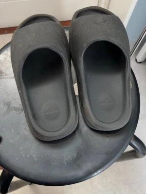 Svarta Adidas tofflor i gummi - Säljer ett par svarta Adidas tofflor i mjukt gummi med öppen tå och bred rem över foten. Perfekta för chill dagar hemma eller till stranden. Klassisk Adidas-logga i sulan och enkel, stilren design.