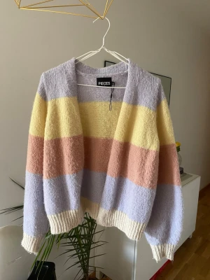 Pastellrandig kofta från Pieces - Mysig kofta från Pieces med breda pastellfärgade ränder i lila, gul och persikorosa. Koftan har öppen front, långa ärmar och ribbade muddar i vitt. Perfekt för dig som gillar en färgglad och avslappnad stil.