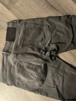 Grå jeans från True Religion - Säljer ett par grå jeans från True Religion med klassiska stora bakfickor och tydliga sömmar. Jeansen har en snygg tvätt och är tillverkade i ett mjukt denimtyg. Perfekta för dig som gillar streetwear och vill ha en unik look.