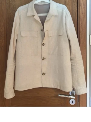 Beige overshirt i linne - Stilren beige overshirt i linne med klassisk krage och fyra knappar framtill. Två stora bröstfickor ger en avslappnad look. Långärmad modell med knapp vid ärmslut. Perfekt för lager-på-lager och enkel att matcha med olika outfits.