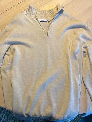 Beige stickad tröja med dragkedja - Snygg beige stickad tröja från RYVLS Stockholm med halv dragkedja och ribbad krage. Tröjan har långa ärmar och ribbade muddar vid ärmslut och nederkant. Perfekt för en stilren och avslappnad look.