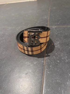 Beige rutigtbälte - Snyggt bälte från med klassiskt beige och brunt rutigt mönster. Bältet har en metallspänne medikoniska TB-logga framtill. Perfekt accessoar för att lyfta din outfit.