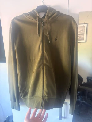 Olivgrön hoodie från Polo Ralph Lauren - Snygg olivgrön hoodie med dragkedja från Polo Ralph Lauren. Klassisk design med huva och dragsko, samt diskret logga broderad på bröstet. Tillverkad i mjukt material och har fickor framtill. Perfekt för en avslappnad och stilren look.