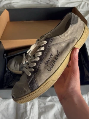 Saint Laurent grå sneakers i canvas - Grå sneakers från Saint Laurent i canvas med beige sula och vita skosnören. Sidan har handskriven logga och skon har en klassisk låg profil. Perfekt för dig som gillar en avslappnad och trendig stil.