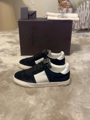 Valentino flycrew  - Snygga sneakers från Valentino i svart mocka med vit läderdetalj på sidan och vit häl. Skorna har svarta skosnören och en vit sula med små nitar på sidan. Perfekta för dig som vill ha en stilren och lyxig look. Skorna är i 9,8/10 i skick det ända är att det saknas en nit men det finns extra nitar, skorna är i storlek 41,5 men sitter som en 42-42,5 