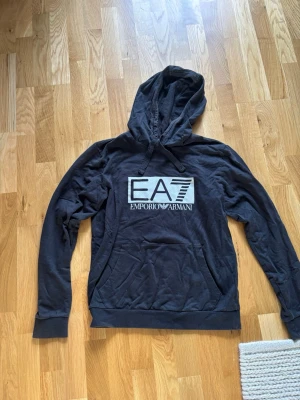 Svart hoodie från Emporio Armani EA7 - Svart hoodie från Emporio Armani EA7 med stor logga på bröstet med luva. Storlek S.