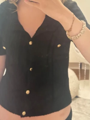 Svart kortärmad blus med guldknappar - Snygg svart blus med korta ärmar och guldfärgade knappar framtill. Modellen är croppad och har en klassisk krage samt dekorativa ficklock med knappar på bröstet. Perfekt för en trendig och stilren look.