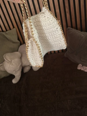 Virkat beige handväska med guldkedja - Snygg handväska i virkat beige garn med guldfärgade kedjehandtag. Väskan har en mjuk, fyrkantig form och känns både trendig och unik. Perfekt accessoar för dig som gillar handgjorda detaljer och en stilren look.