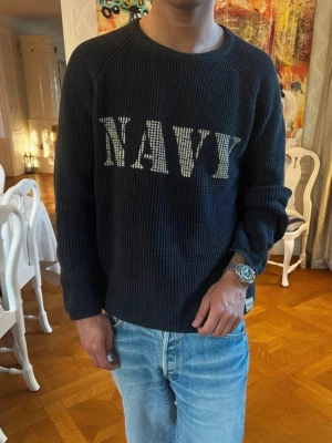 Waffle knit Navy Pullover vintage - Säljer en mörkblå stickad tröja med vit text 'NAVY' framtill. Tröjan har rund halsringning och avslappnad passform. Perfekt för dig som gillar en casual och cool stil. Materialet ser ut att vara mjukt och bekvämt, och tröjan har långa ärmar.
