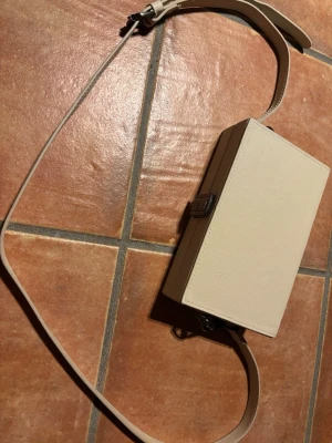 Beige boxväska från Zara - Stilren beige handväska från Zara i boxmodell med justerbar axelrem. Väskan har en hård rektangulär form, metallspänne och svart tygfoder på insidan. Perfekt för dig som gillar minimalistisk design och vill ha en unik accessoar.