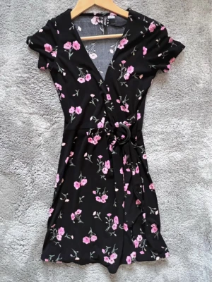 Svart blommig omlottklänning - Kort svart omlottklänning med rosa blommor och knytbälte med rund spänne i midjan. Klänningen har v-ringning och korta ärmar, perfekt för en trendig och feminin look. Materialet känns mjukt och följsamt.