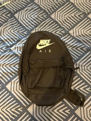 Svart ryggsäck Nike Air - Snygg svart ryggsäck från Nike Air med klassisk logga i vitt framtill. Ryggsäcken har ett stort huvudfack, en mindre ficka framtill och justerbara, vadderade axelband. Tillverkad i slitstarkt material, perfekt för skolan eller träningen.