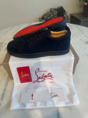 Mörkblå Christian Louboutin sneakers - Snygga mörkblå sneakers från Christian Louboutin med klassisk röd sula och diskret logga. Skorna är låga och har ovandel i mocka med matchande mörkblå snörning. Perfekta för dig som vill ha en exklusiv och stilren look.