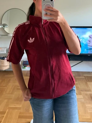 Vinröd Adidas zip-tröja kort ärm - Säljer en vinröd Adidas zip-tröja med korta ärmar och vit logga på ärmarna.Jättesnygg men kommer inte till någon användning. Är egentligen i stl 3 Xl men jag har vanligtvis stl M och den sitter snyggt! Har vikt upp nederdelen lite men inget som syns (fråga om du vill ha fler bilder på det) Sitter jättesnyggt men har tröttnat på den:)