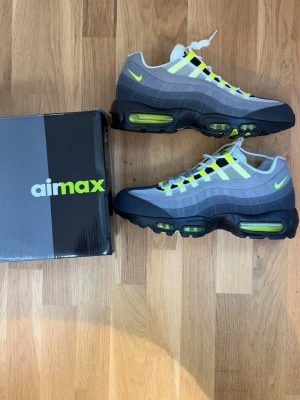 Nike Air Max 95 Neon sneakers - Nike Air Max 95 sneakers i grå, svart och vit med neongula detaljer. Klassisk vågig design, synliga Air-enheter i sulan och meshpaneler för bra ventilation. Ikonisk logga på plös och häl. Perfekta för dig som vill sticka ut med stilrena och sportiga sneakers.