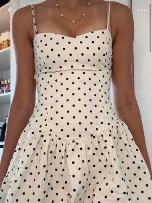 Vit prickig klänning från Bershka - Supersöt vit klänning från Bershka med svarta prickar och smala axelband. Klänningen har en figurnära överdel och utställd kjol som ger en lekfull look. Perfekt för varma dagar och somriga vibes!