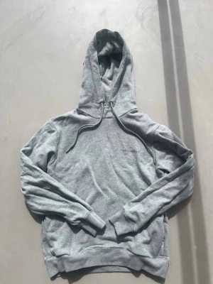Ljusgrå hoodie med luva - Tiger of Sweden - En stilren ljusgrå hoodie med huva och dragsnören. Hoodien har ribbade muddar vid ärmslut och nederkant samt en klassisk passform. Tillverkad i mjukt sweatshirtmaterial, perfekt för en avslappnad look.