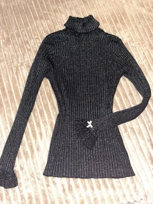Svart glittrig polo blus - Säljer en svart glittrig ribbstickad polo blus. Tröjan har lång ärm och figurnära passform, en liten rosa rosett på armarna och en liten öppning vid nacken. 