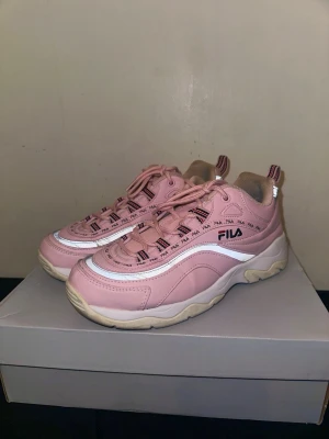 Rosa Fila sneakers med Reflex - Snygga rosa sneakers från Fila med vit och beige chunky sula. Skorna har vita och svarta detaljer samt Fila-logga på sidan och längs snörningen. Ovandelen är i syntetmaterial och textil, med dragflikar bak och fram för enkel påtagning.