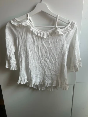 Vit volangtopp med trekvartsärm - Söt vit topp med volangdetaljer vid halsringning, ärmslut och nederkant. Toppen har trekvartsärmar och offshoulder-design med smala axelband. Perfekt för en trendig och feminin look.
