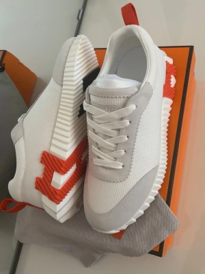 H skor - Snygga vita H sneakers med ribbad sula och kontrasterande röd och svart H-detalj på sidan. Ovandelen är i textil och mocka, med vita skosnören och röd hälflik. Perfekta för dig som gillar exklusiv och modern stil.