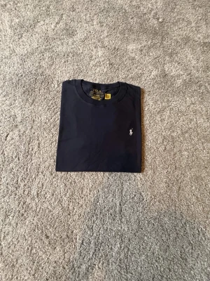 Ralph lauren T-shirt mörkblå Ny-oanvänd (M) -  Säljer en helt ny och oanvänd Ralph Lauren T-shirt i storlek M. Stilren och tidlös T-shirt i mörkblå färg med den klassiska vita Ralph Lauren-loggan broderad på bröstet. Tillverkad i mjukt och bekvämt material av hög kvalitet – perfekt för både vardag och en avslappnad, stilren look. 🔹 Märke: Ralph Lauren 🔹 Modell: T-shirt 🔹 Färg: Mörkblå med vit logga 🔹 Storlek: M 🔹 Skick: Helt ny / oanvänd 🔹 Passform: Klassisk Nypris är högt – perfekt möjlighet till ett fynd
