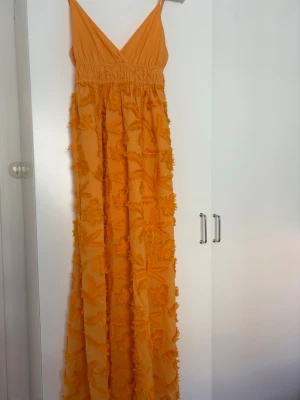 Lång orange klänning  - Säljer en lång, orange klänning med v-ringning och smala axelband. Klänningen har ett blommigt mönster i struktur och är tillverkad i ett lätt, luftigt material som passar perfekt till sommaren. Den har en markerad midja och faller snyggt längs kroppen.