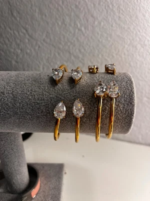 Guldfärgat armband med droppformade stenar - Säljs styckvis!                                            Snyggt armband i guldfärgad metall med två stora, droppformade klara stenar i varje ände. Armbandet har en öppen design och ger en lyxig känsla med sina glittrande detaljer. Perfekt accessoar för att lyfta din outfit.