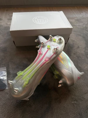 Adidas F50 Elite silver fotbollsskor - Säljer ett par Adidas F50 fotbollsskor i silver med neonrosa och gula detaljer. Skorna har metall-dobbar och en strömlinjeformad design utan snörning för maxad speed på planen. Tillverkade i syntetmaterial för lätt känsla och bra grepp. Perfekta för dig som vill sticka ut på matchen.