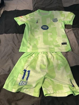 Barcelona fotbollskit Raphinha #11 - Säljer ett limegrönt Barcelona matchställ från Nike med Raphinha och nummer 11 på ryggen. Setet består av en kortärmad tröja och shorts med klubbmärke, sponsorloggor och tryck i blått. Perfekt för dig som älskar fotboll och vill sticka ut på planen.