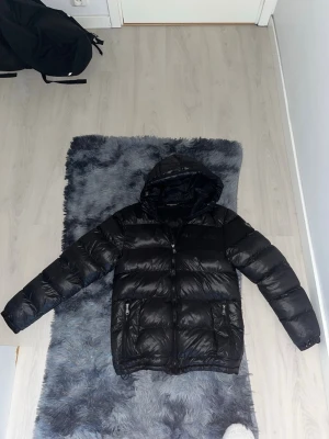 Alexandro zavetti pufferjacka - Säljer en svart pufferjacka med huva och dragkedja framtill. Jackan har två fickor med dragkedja och quiltad design som ger en riktigt fet look. Perfekt för kalla dagar när du vill hålla värmen och ändå se stilren ut.